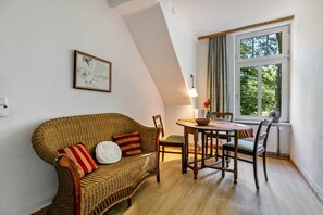 Ein Zimmer mit einem Korbsessel, einem Esstisch, Stühlen, einer Lampe, einem gerahmten Bild und einem Fenster mit Vorhängen.