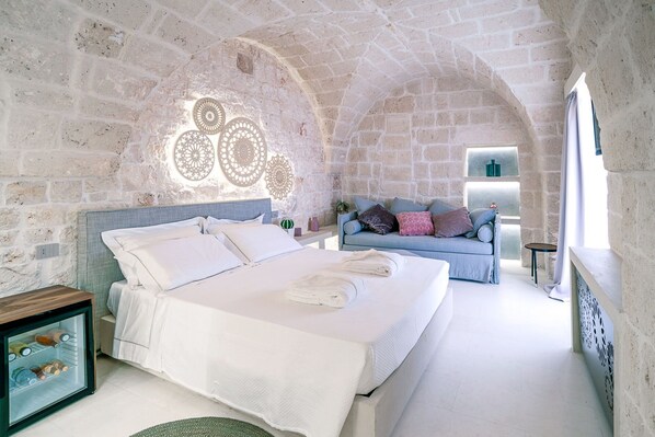 A bedroom with a stone wall, a bed with white linens, a blue sofa with cushions, and a small table with a plant.