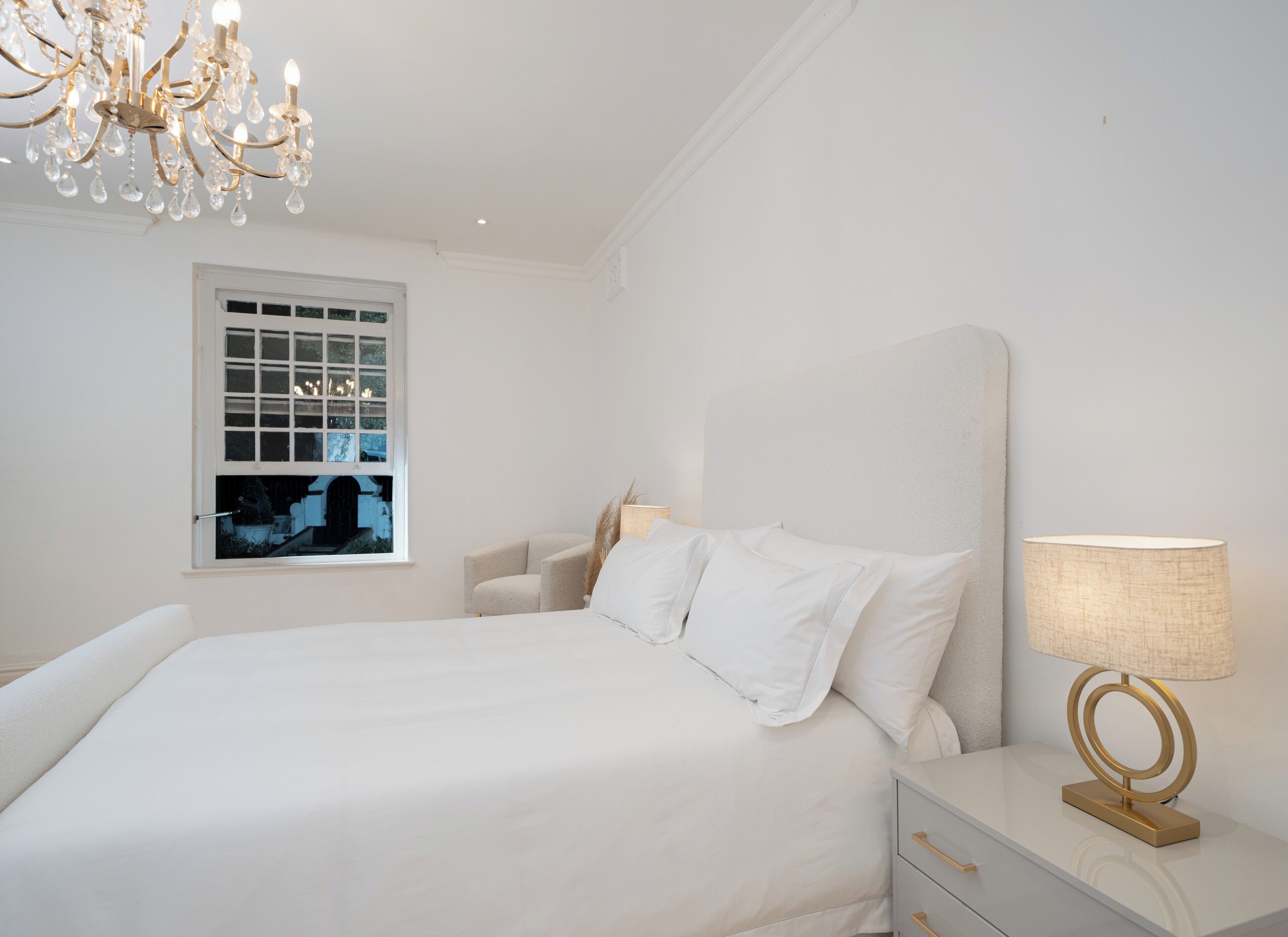 Un dormitorio blanco con una cama grande, una mesita de noche con lámpara y una ventana con vista a un edificio.