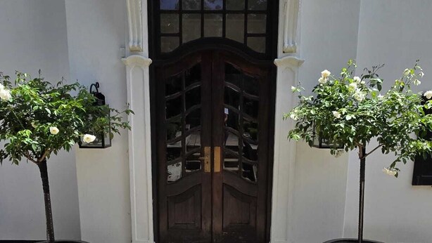 Entrada de un edificio con dos puertas de madera oscura, flanqueada por dos grandes macetas con árboles de flores blancas.