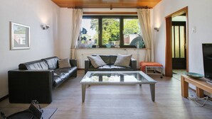 Een woonkamer met een glazen salontafel, een zwarte leren bank, een houten bankje en een televisie op een houten dressoir.