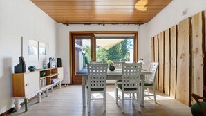 Een eetruimte met een houten plafond, witte stoelen met groene kussens, een houten tafel en een raam met uitzicht op groen.