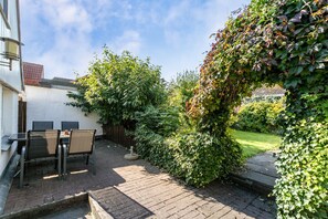 Een terras met een tafel en stoelen, omgeven door groen en een witte muur.