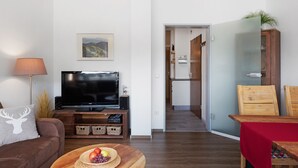 Un salon avec un téléviseur à écran plat, un canapé orné d’un coussin à motif de cerf, une table basse en bois sur laquelle sont disposés des fruits, et un coin cuisine à l’arrière-plan.