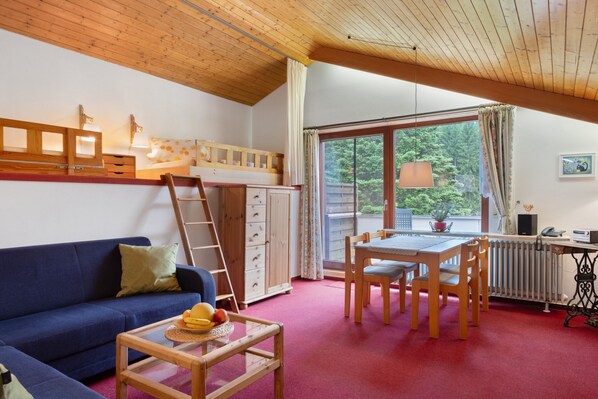 Een gezellige kamer met een blauwe bank, een houten tafel en een zithoek met stoelen. Een ladder staat tegen een garderobe en een radiator is zichtbaar.