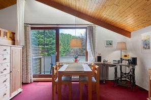 A room with a dining table, chairs, a radiator, a sewing machine, and a window with a view of trees. - Apartment 1 'Eule, 42qm' with Balcony and Wi-Fi (Schönwald im Schwarzwald)