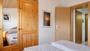 A bedroom with a wooden wardrobe, a bed with checkered bedding, a framed picture of a sunset, and a door leading to another room.