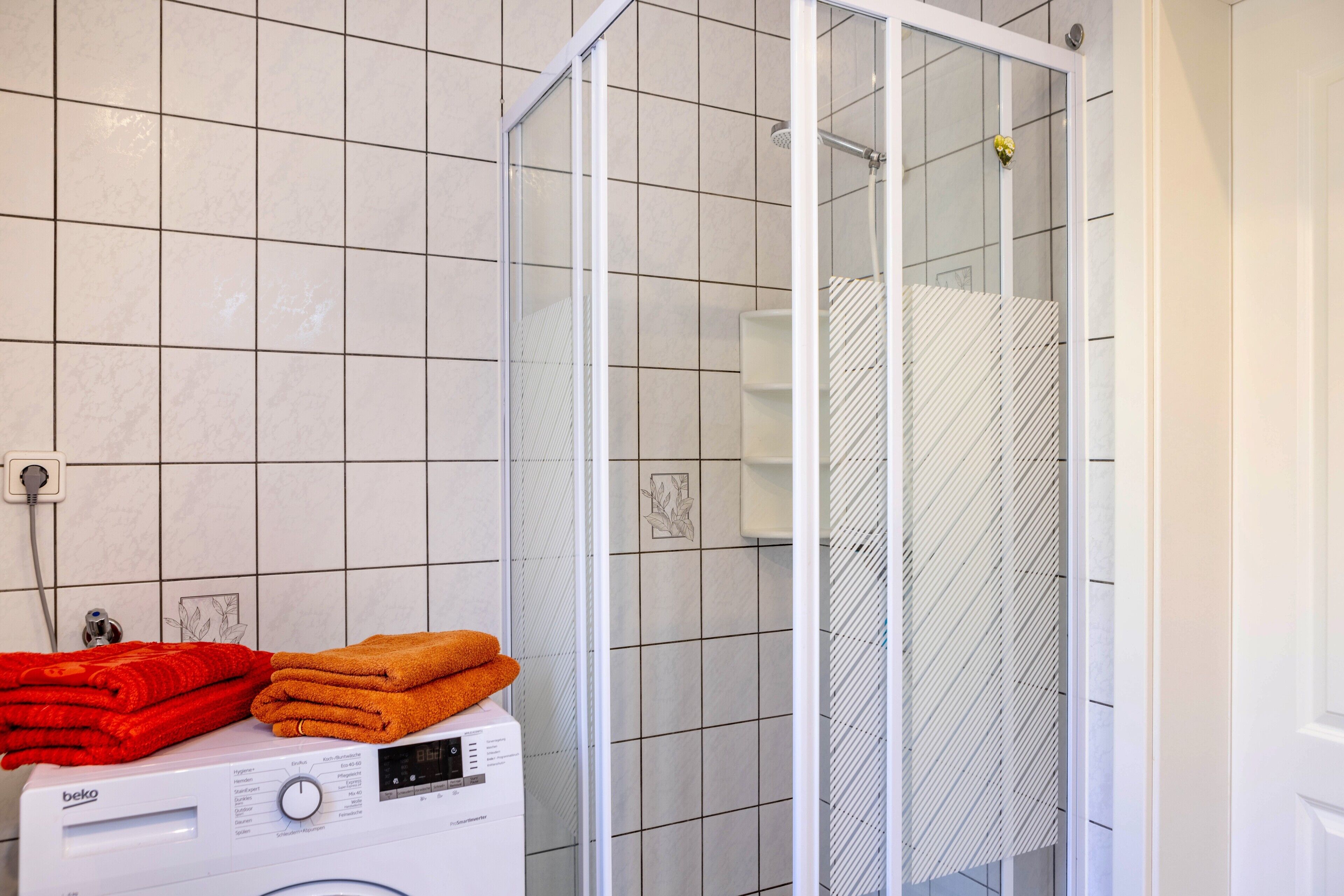 Un baño con lavadora, cabina de ducha de vidrio y paredes de azulejos.