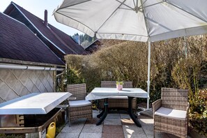 En udendørs terrasse med et bord og stole, en hvid parasol og en bygning med mørkt tag i baggrunden.