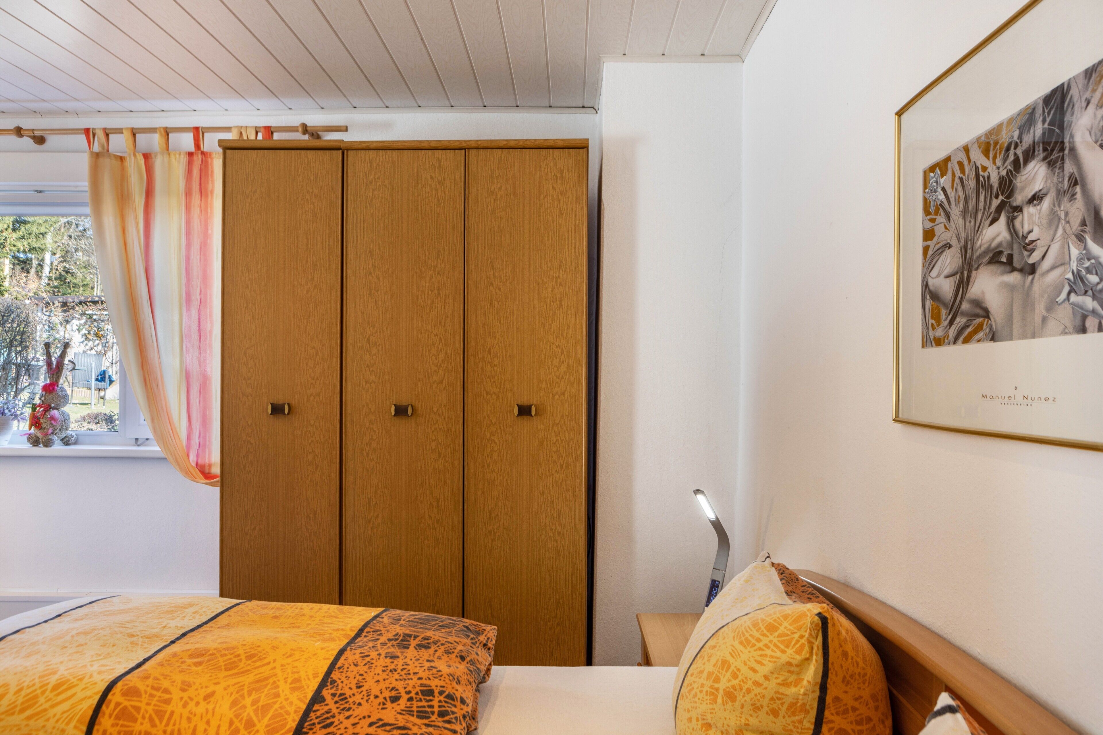 A bedroom with a bed, wooden wardrobe, and a framed artwork on the wall.
