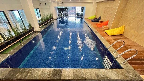 A large indoor swimming pool with a clear blue water surface, surrounded by wooden flooring and colorful cushions. - Front One Boutique Adria Boyolali (Boyolali)