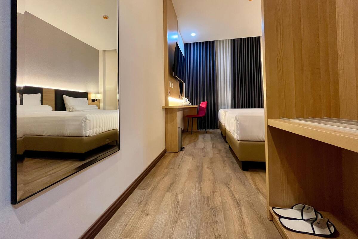 Une chambre d’hôtel avec un grand miroir, deux lits, un bureau avec une chaise, une télévision et une paire de chaussons posés sur le sol.