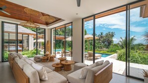 A modern living room with a large sofa, wooden accents, and a view of a pool and garden. - Rosita Ubud - Stunning 5BR Villa  (Ubud)