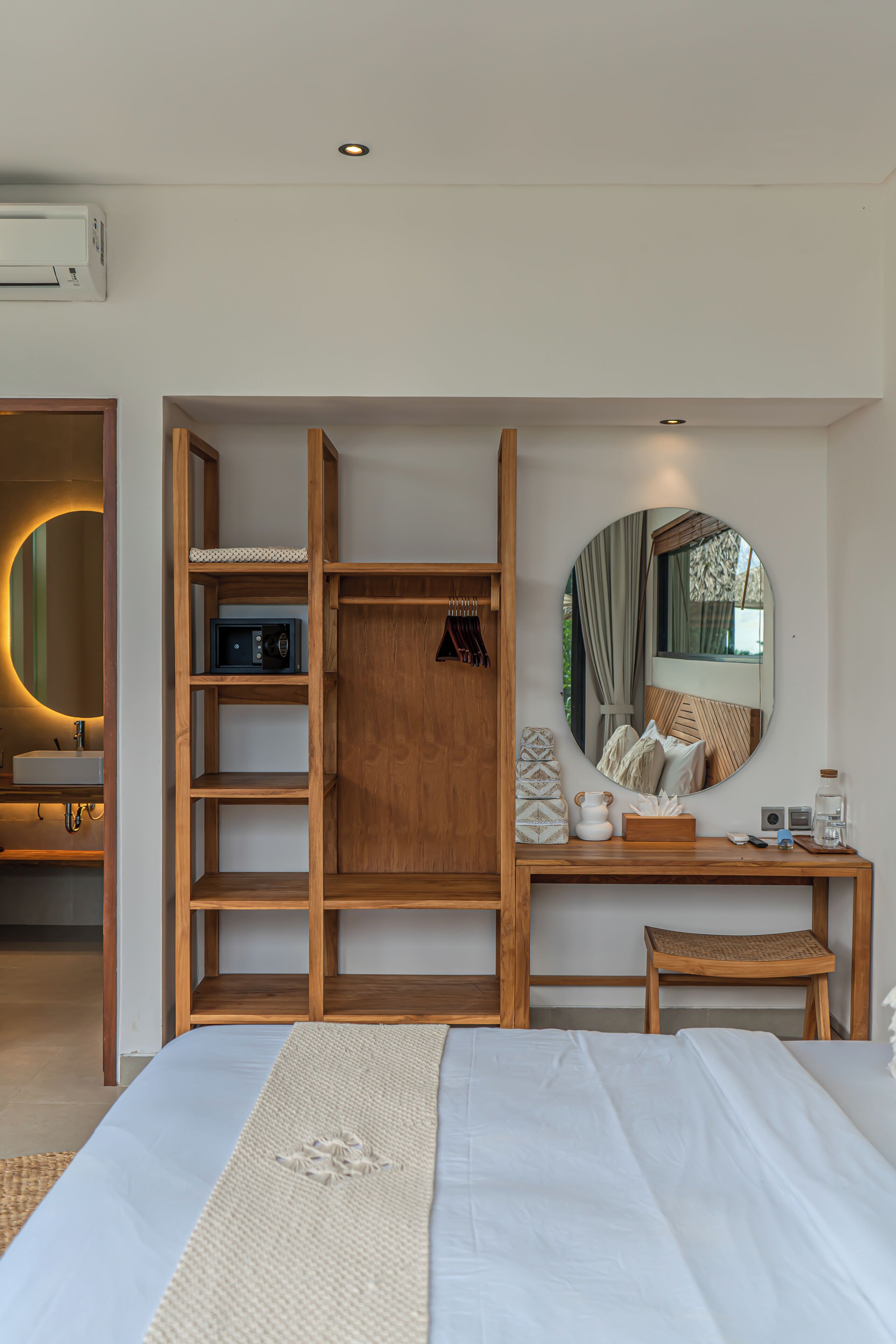 Un dormitorio con una cama, un armario de madera, un espejo redondo, una mesita de noche y un taburete pequeño.