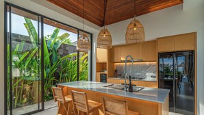 A modern kitchen with a large island, wooden chairs, and a view of lush greenery through large windows. - Rosita Ubud - Stunning 5BR Villa  (Ubud)