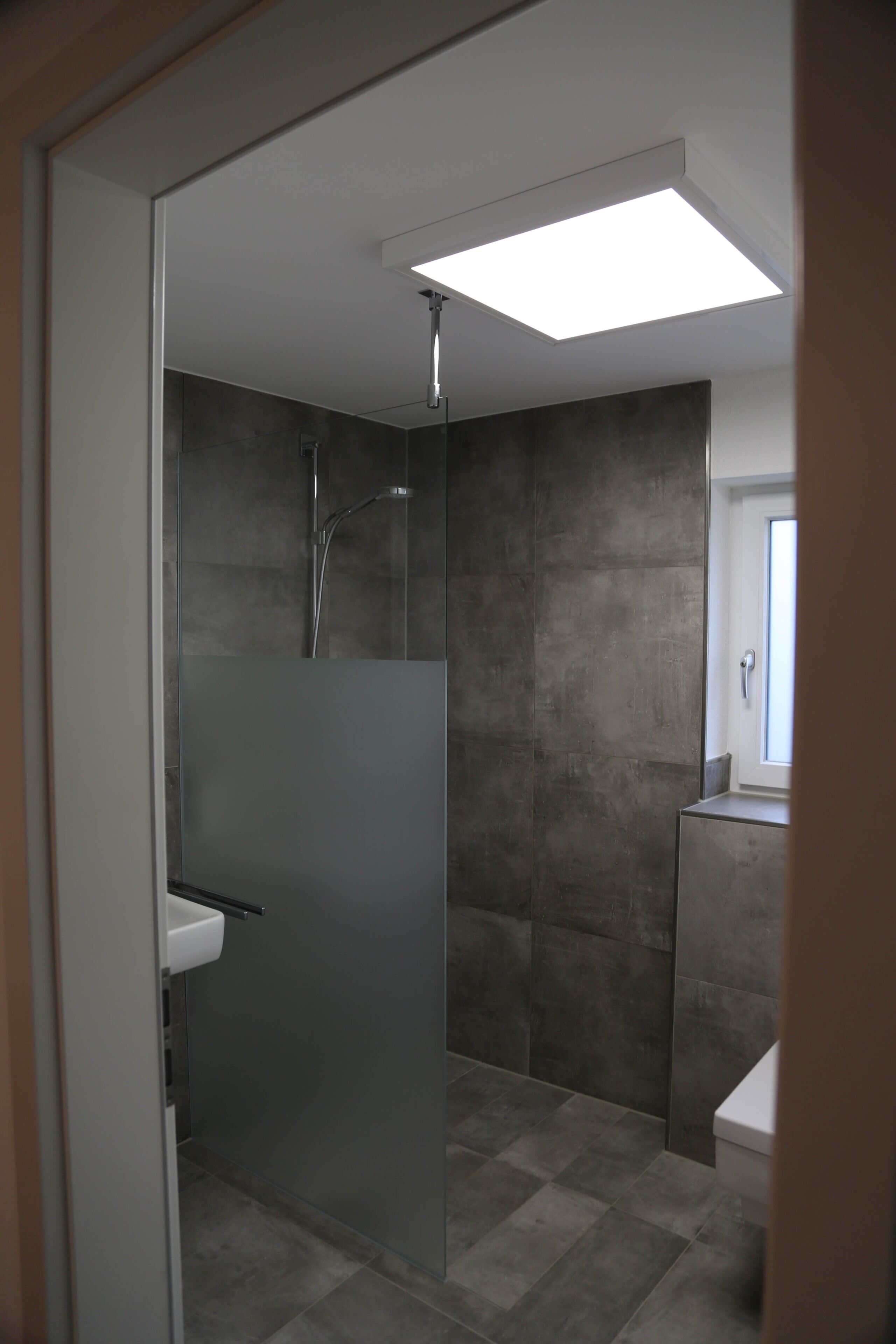 Une salle de bain moderne dotée d’une douche à parois de verre, d’un lavabo blanc et d’une grande fenêtre qui laisse entrer la lumière naturelle.
