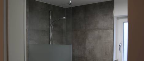 Une salle de bain moderne dotée d’une douche à parois de verre, d’un lavabo blanc et d’une grande fenêtre qui laisse entrer la lumière naturelle.