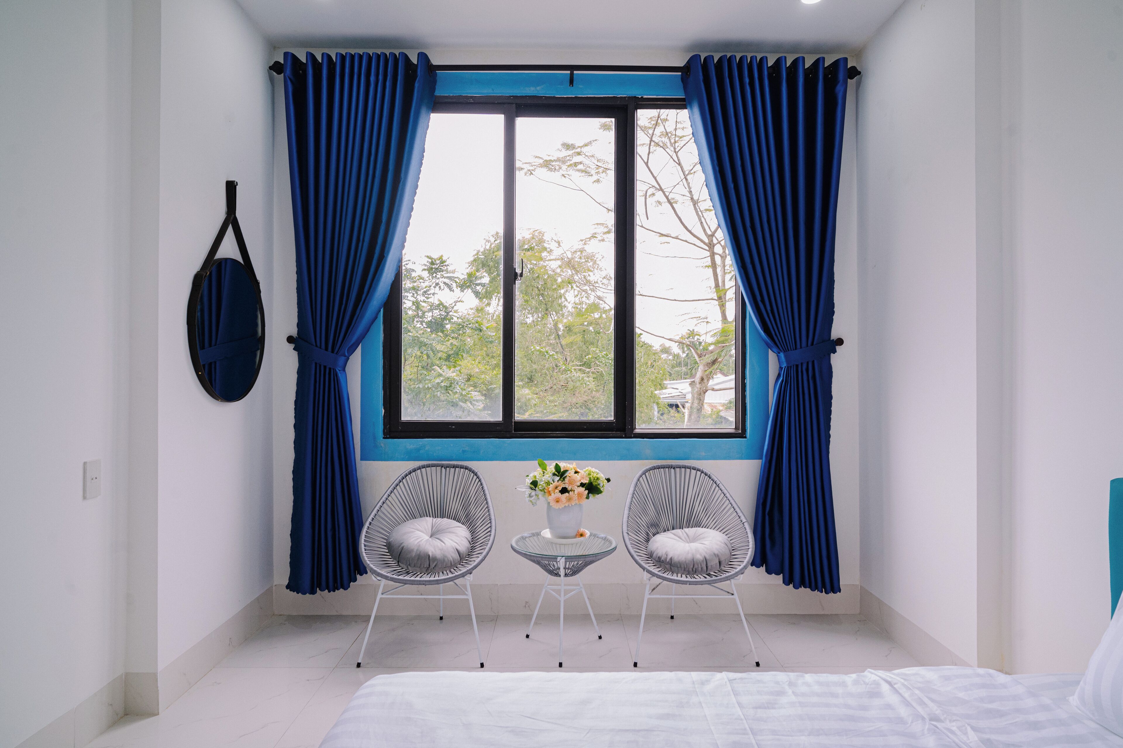 A bedroom with a bed, two chairs, a small table with a vase, and a window with curtains.