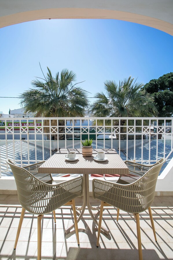 A terrace with a table and chairs, a potted plant, and a view of the sea and palm trees.
