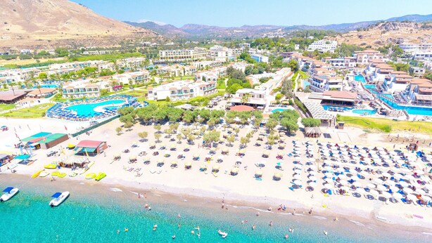 Vista aérea de un complejo turístico costero con varias edificaciones, una gran piscina y una playa con tumbonas.