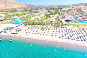 An aerial view of a coastal resort with multiple buildings, a large swimming pool, and a beach with sun loungers.