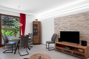 A modern living room with a stone wall, a flat-screen TV, a wooden cabinet, and a dining area with chairs and a table.
