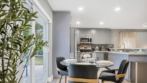Una cocina moderna con electrodomésticos de acero inoxidable, un comedor con mesa y sillas de vidrio, y vistas al exterior.