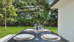 Mesa al aire libre con platos, vasos y un florero, rodeada de césped y árboles.