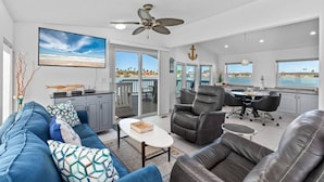 A spacious living area with a large TV, a ceiling fan, a dining table with chairs, and a view of the ocean. - Luxury 168 Long Island Village (Port Isabel)