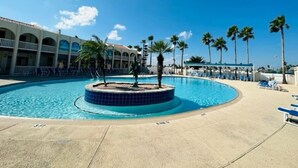 A swimming pool with a tiled center, surrounded by lounge chairs and palm trees, with a building in the background. - Luxury 168 Long Island Village (Port Isabel)