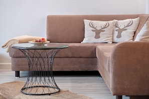A brown sofa with two deer head pillows, a glass coffee table with a woven base, and a plate of fruit on the sofa.