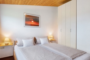 A bedroom with a bed, two bedside tables with lamps, a wardrobe, and a framed sunset picture on the wall.