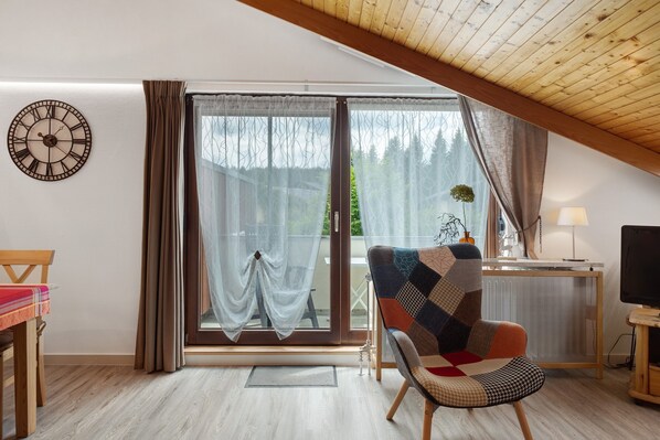 A room with a wooden ceiling, a large window with curtains, a clock on the wall, a chair, a table, and a TV.