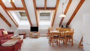 Ein Zimmer mit schrägem Dach, Oberlichtern, einer Couch, einem Esstisch, Stühlen, einem Fernseher und einer Heizung.