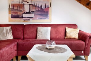 Un divano rosso con tre cuscini, un vaso di vetro su un tavolo bianco e un quadro appeso al muro.