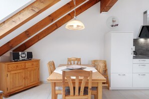 Un coin repas comprenant une table en bois, des chaises, un luminaire suspendu, un meuble blanc et un espace cuisine avec une cuisinière.
