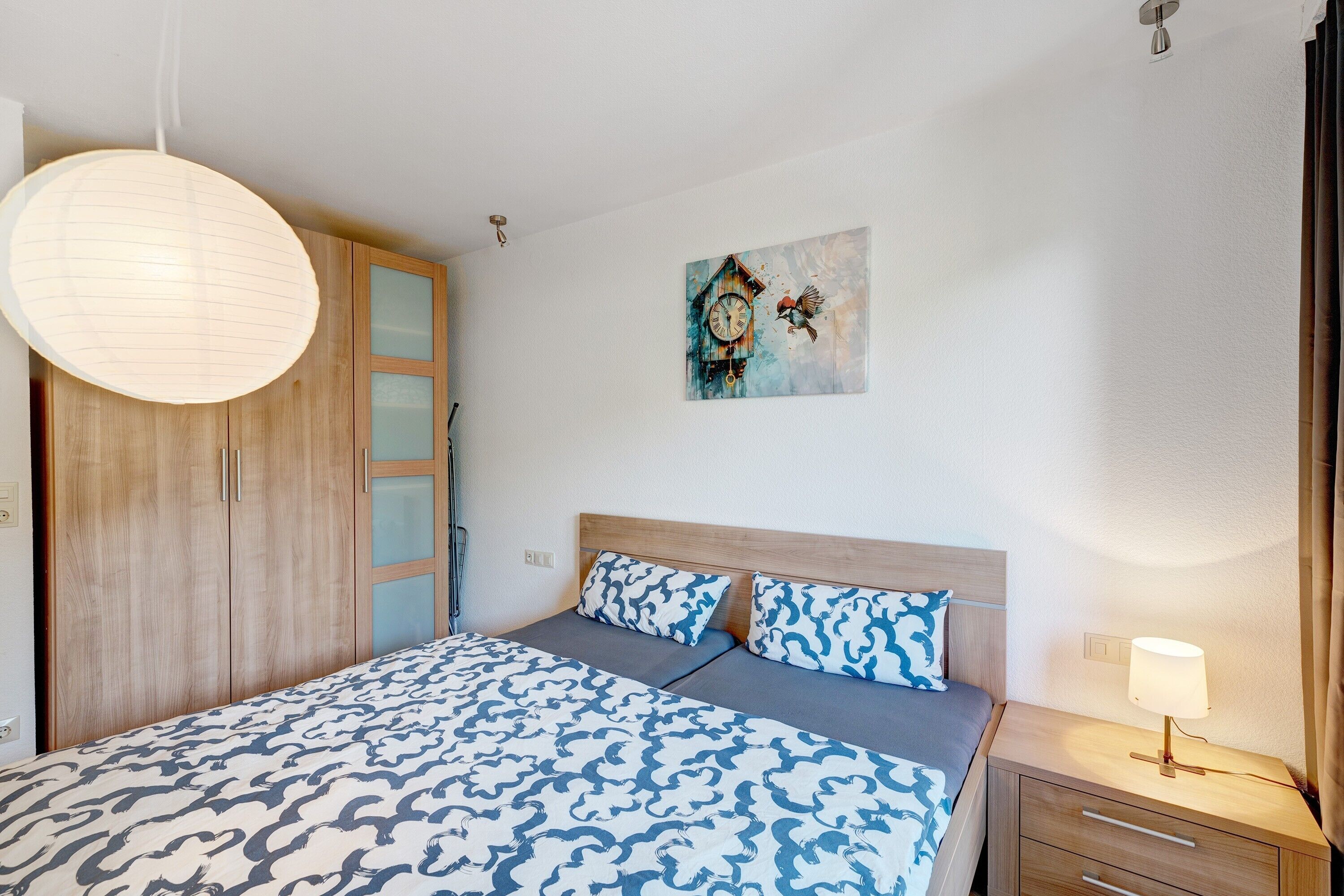 A bedroom with a bed, a wooden wardrobe, a nightstand, a wall clock, and a hanging light fixture.