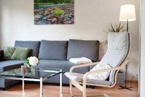 A modern living room with a glass coffee table, a grey sofa, a wooden chair with a cushion, a floor lamp, and a painting on the wall. - Apartment 'Am Lehen' with Mountain View, Private Terrace and Wi-Fi (Schiltach)