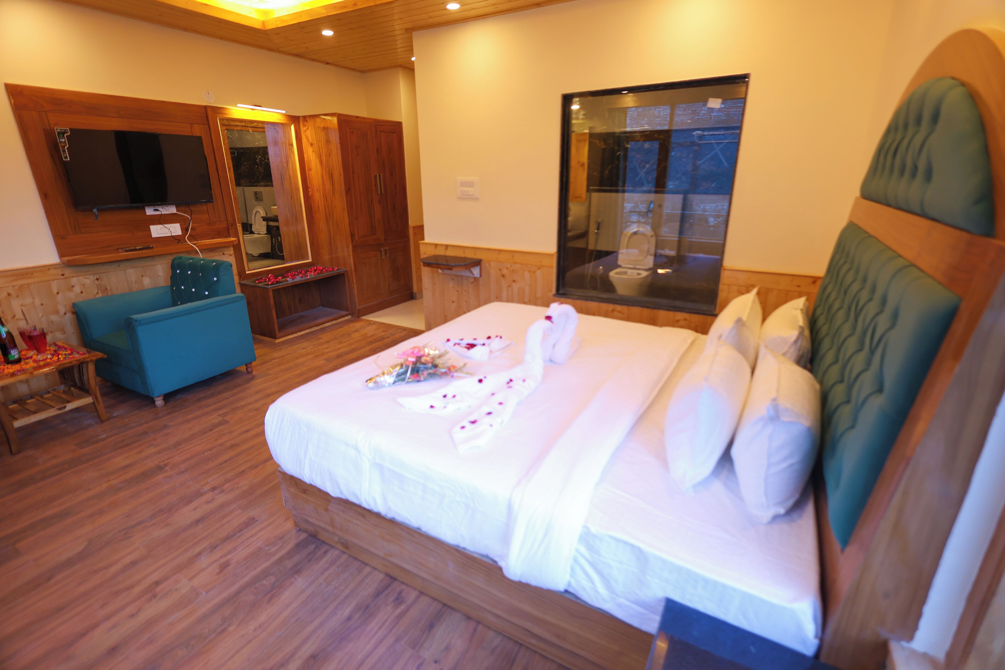 Un dormitorio con una cama grande, cabecera de madera, un sillón azul, un armario de madera, un televisor y un baño con mampara de vidrio.
