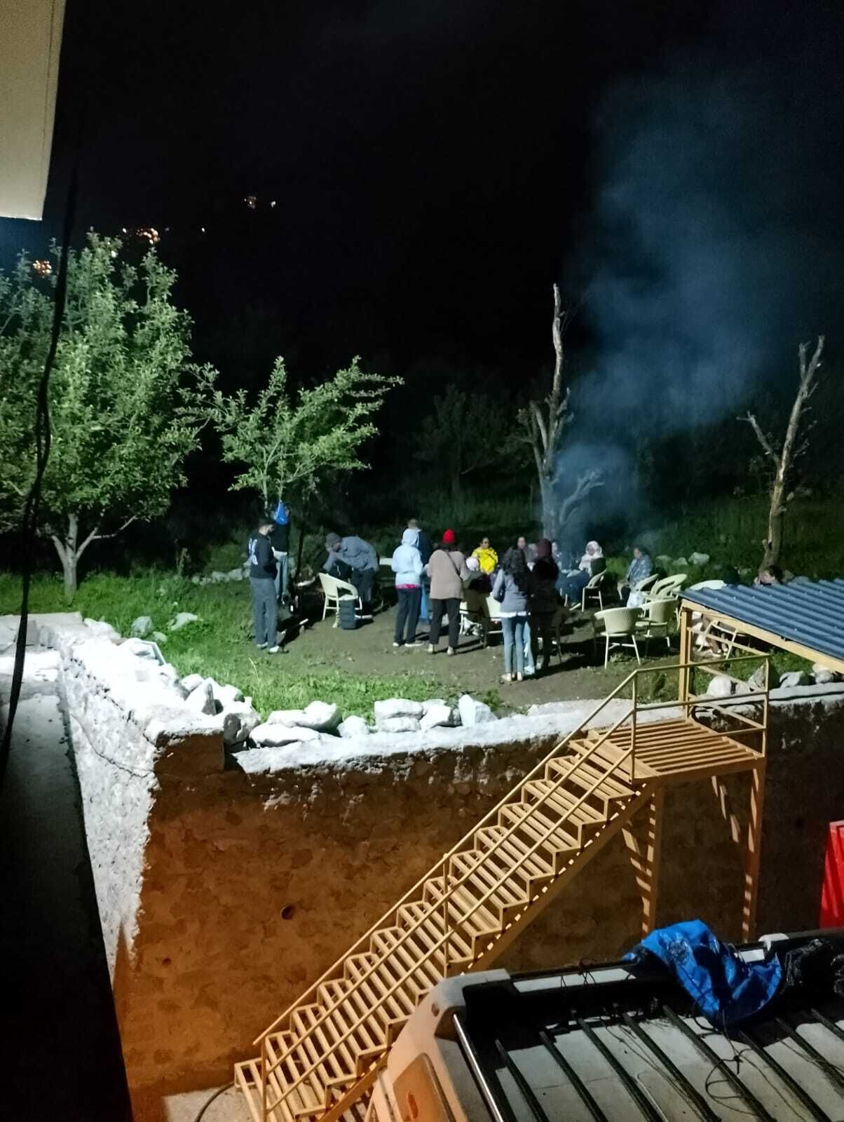 Un groupe de personnes s’est réuni à l’extérieur, de nuit, autour d’un feu de camp, avec une structure en bois en arrière-plan.