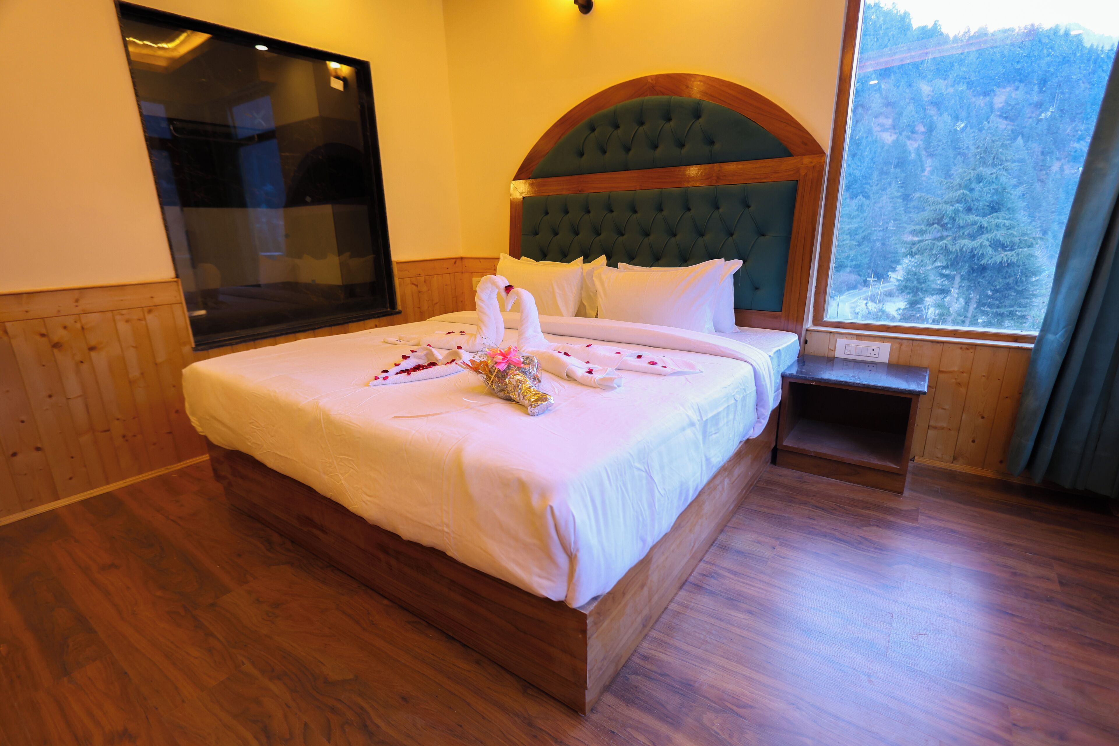 A neatly made bed with a wooden headboard, a large window with a view of trees, and a wooden floor.