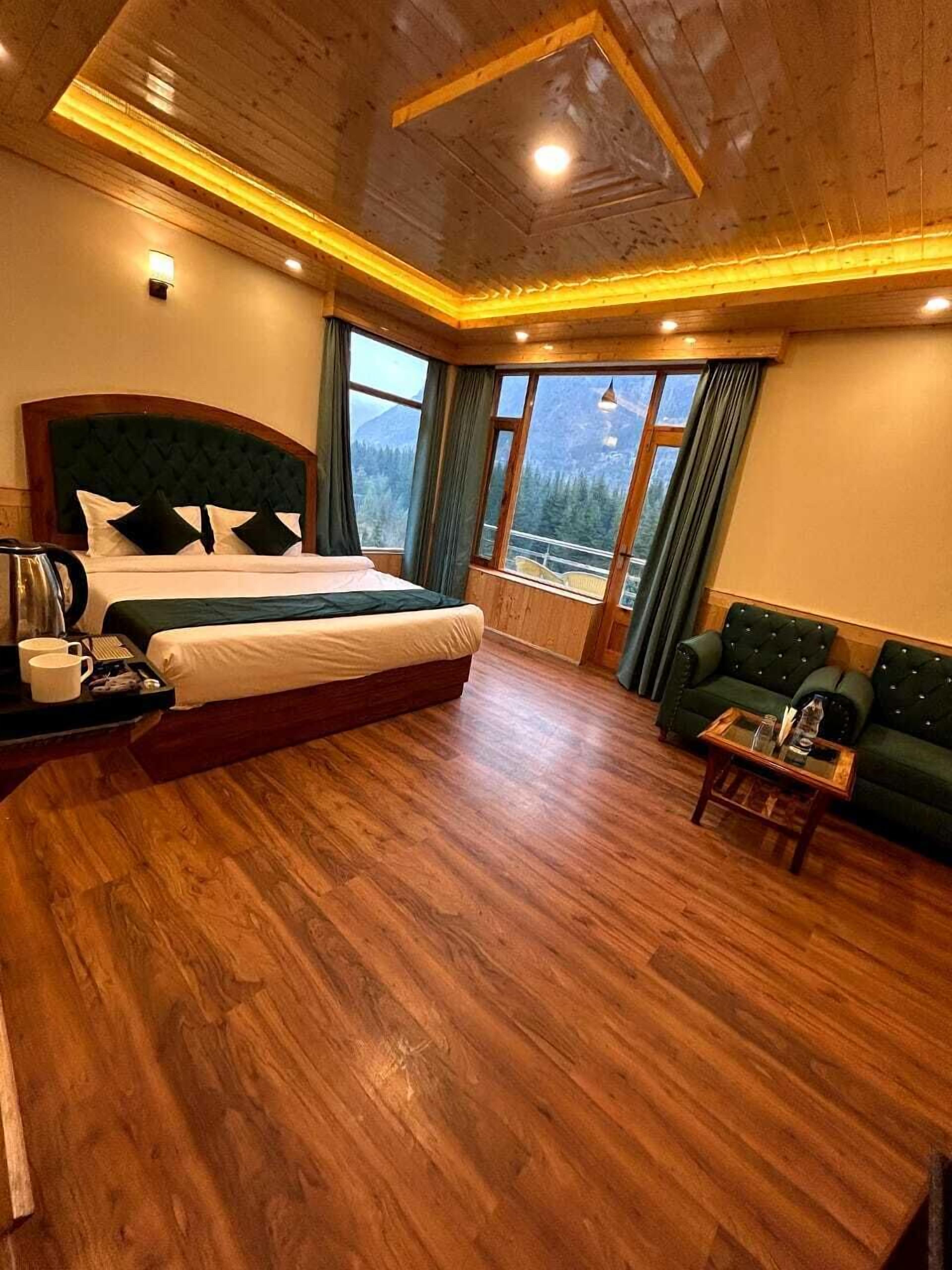 A bedroom with a large bed, a green sofa, wooden flooring, and a view of mountains through the windows.
