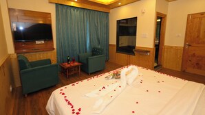 Una habitación de hotel con una cama grande, televisión, un sillón verde, una mesita con una botella y vasos, y techo de madera.