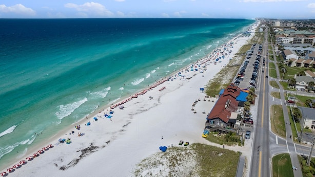 An aerial view of a beach with rows of umbrellas and beach chairs, a road parallel to the beach, and buildings along the shore. - Sleeps 18! Private Pool, Hot Tub, Near Beach | Miramar Dream House by AvantStay (Miramar Beach)