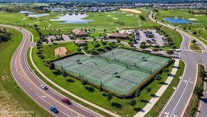 An aerial view of a golf course with a large tennis court, multiple buildings, and a road curving around the area.