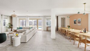 Un salon spacieux avec un grand canapé, un coin repas avec une longue table et des chaises, et une vue sur l’extérieur grâce à de grandes fenêtres.