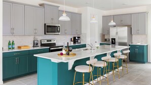 Une cuisine moderne avec des meubles de couleur turquoise, un îlot central blanc doté de tabourets, des appareils en acier inoxydable et des luminaires suspendus.