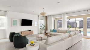 Un salon spacieux avec un grand canapé d’angle beige, une table basse blanche et un pouf vert. Un téléviseur à écran plat est fixé au mur, ainsi qu’une plante en pot. De grandes fenêtres offrent une vue sur une piscine.