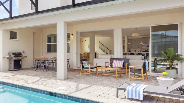 Un espace aménagé au bord de la piscine, comprenant un barbecue, des sièges d’extérieur et une vue sur l’intérieur de la maison.