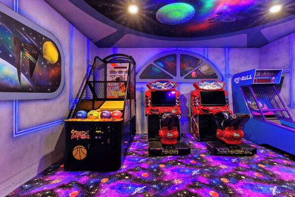 An arcade room with basketball and racing game machines, a basketball hoop, and a space-themed mural. - On-Site Waterpark, Pool & Sleeps 26 | Resort Home – Flamingo Grove by AvantStay (Davenport)